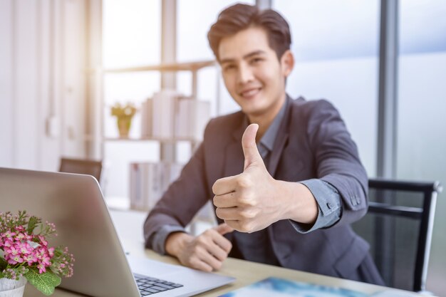 Il giovane uomo d'affari asiatico vede un business plan di successo sul computer portatile e sulla carta per documenti Messa a fuoco sfocata astratta che mostra con i pollici in su sullo sfondo del tavolo in legno in ufficio