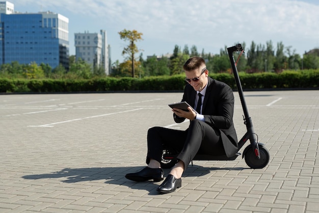 Il giovane uomo d'affari bello in un vestito è seduto su uno scooter elettrico e sta guardando un computer portatile