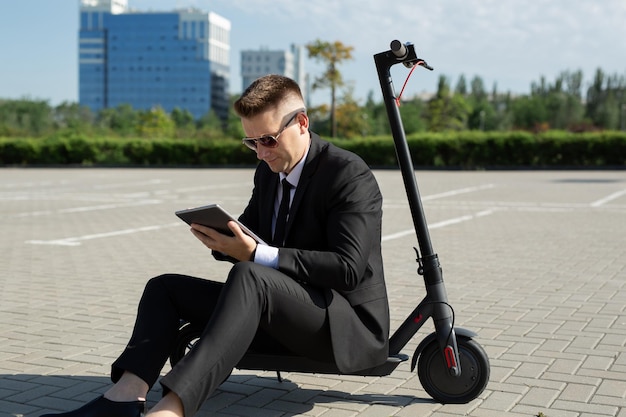 Il giovane uomo d'affari bello in un vestito è seduto su uno scooter elettrico e sta guardando un computer portatile