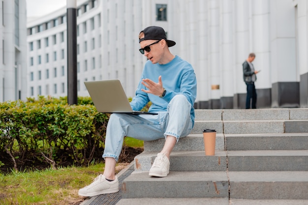 Il giovane uomo d'affari di moda hipster con occhiali da sole si siede sulle scale del centro affari, con una tazza di caffè e usa il laptop