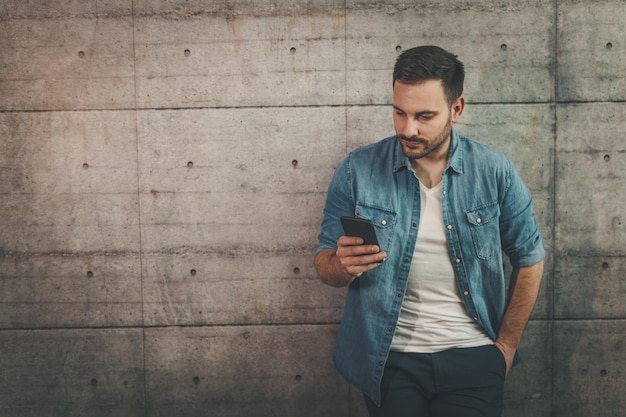 Il giovane uomo d'affari di successo sta leggendo i messaggi su uno smartphone davanti a un muro di cemento.