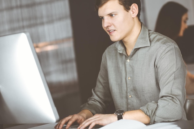 Il giovane uomo d'affari e programmatore dai capelli scuri in una camicia verde sta lavorando sodo sul suo computer, mentre è seduto alla scrivania in un moderno armadietto con una collega sullo sfondo. Concetto di successo