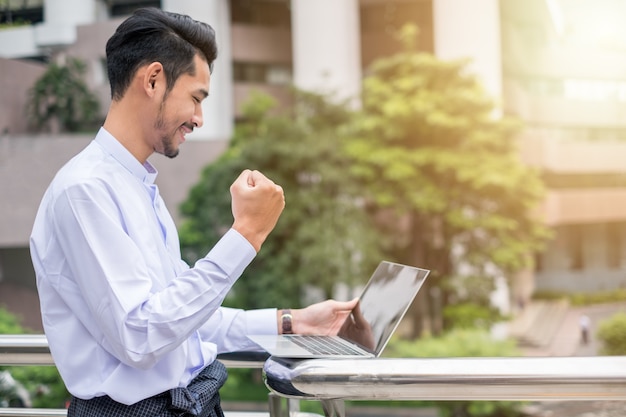 Il giovane uomo d'affari eccitato alza la mano sii felice e sorride con il computer portatile a urban