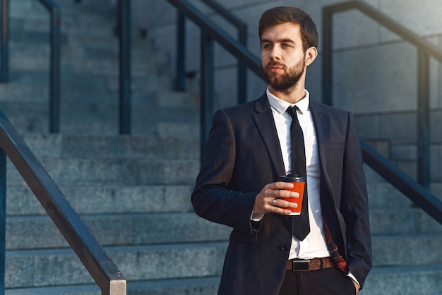 Il giovane uomo d&#39;affari in un vestito che tiene uno sguardo di tazza di caffè di carta rosso al lato.