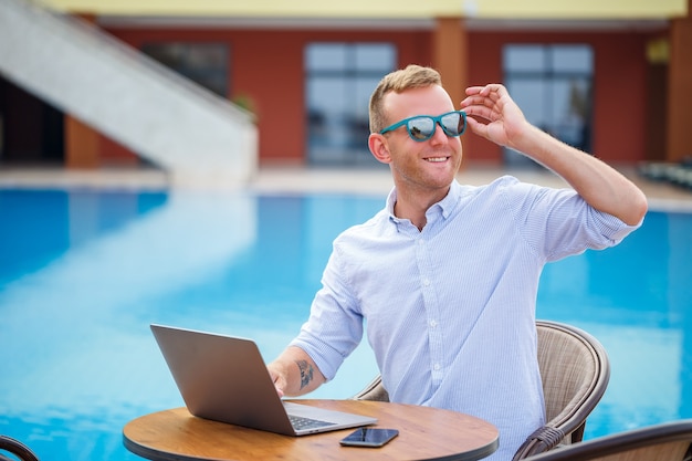 Il giovane uomo d'affari maschio in occhiali da sole lavora a un computer portatile seduto a un tavolo vicino alla piscina. Lavoro a distanza. Libero professionista