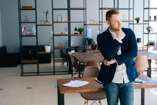 Il giovane uomo d'affari pensieroso che indossa un abito alla moda è in piedi nella moderna stanza dell'ufficio vicino alla scrivania in legno Concetto di lavoro d'ufficio