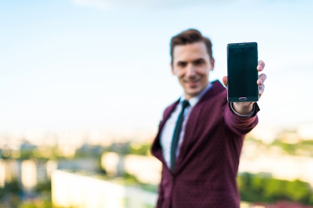 Il giovane uomo d'affari serio in vestito e camicia rossi con il legame sta sul tetto e mostra il telefono vuoto