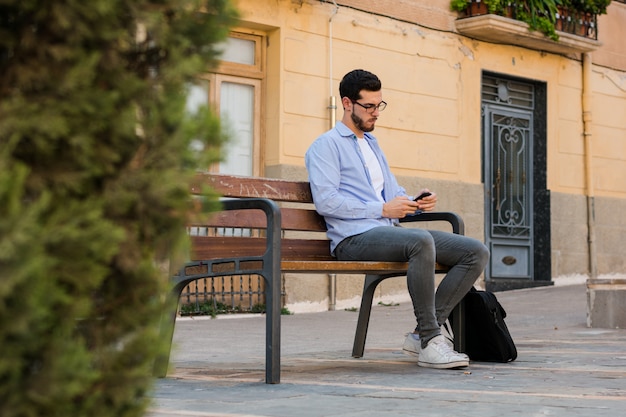 Il giovane uomo d'affari sta sedendosi su una panchina mentre sta parlando sul cellulare