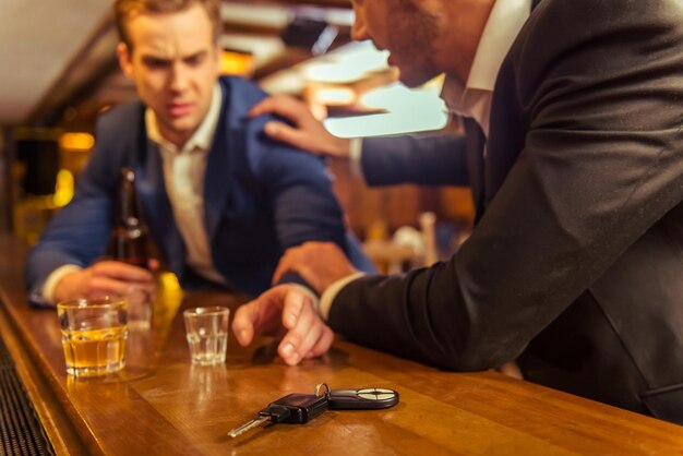 Il giovane uomo d'affari ubriaco tiene in mano una bottiglia di birra e raggiunge le chiavi della macchina mentre è seduto al bancone del bar nel pub un altro uomo lo sta fermando