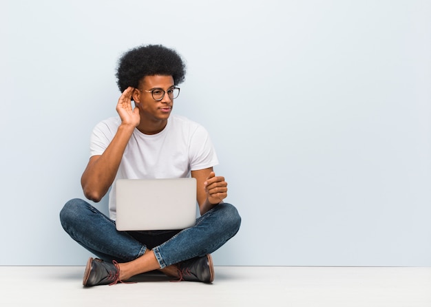 Il giovane uomo di colore che si siede sul pavimento con un computer portatile prova ad ascoltare un gossip