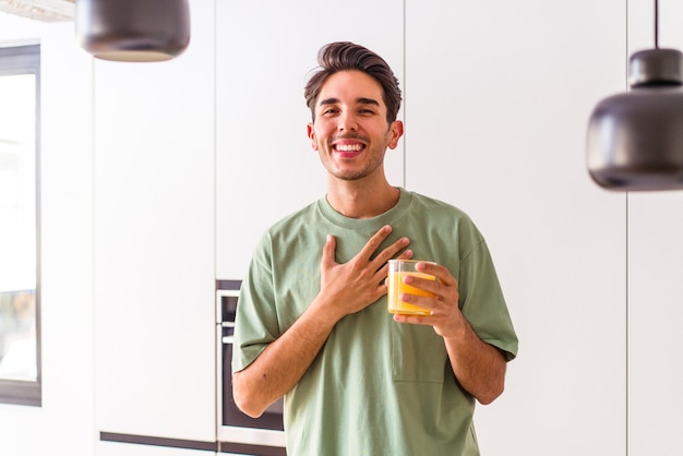Il giovane uomo di razza mista che beve succo d'arancia nella sua cucina ride forte tenendo la mano sul petto.