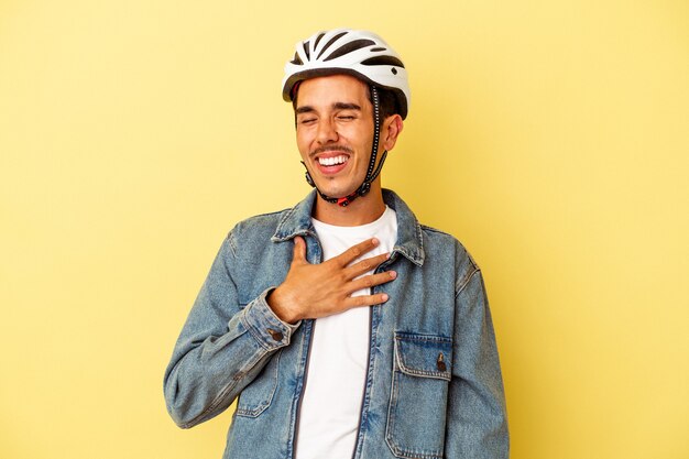 Il giovane uomo di razza mista che indossa un casco da bici isolato su sfondo giallo ride ad alta voce tenendo la mano sul petto.