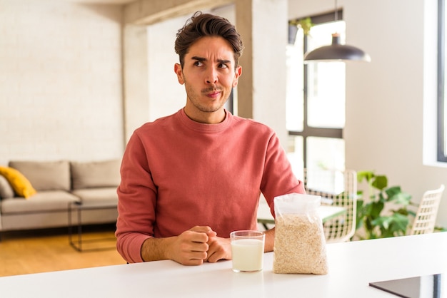 Il giovane uomo di razza mista che mangia farina d'avena e latte a colazione nella sua cucina confuso, si sente dubbioso e insicuro.