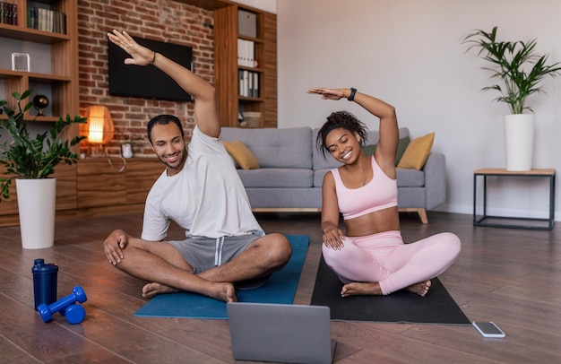 Il giovane uomo e la signora afroamericani sorridenti in abbigliamento sportivo fanno allungamento della mano praticando lo yoga guarda la video lezione