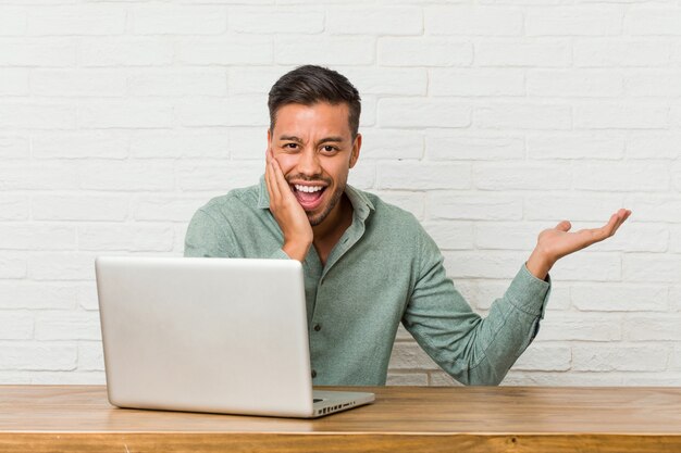 Il giovane uomo filippino seduto a lavorare con il suo laptop tiene lo spazio della copia su un palmo, tenere la mano sulla guancia. Stupito e deliziato.