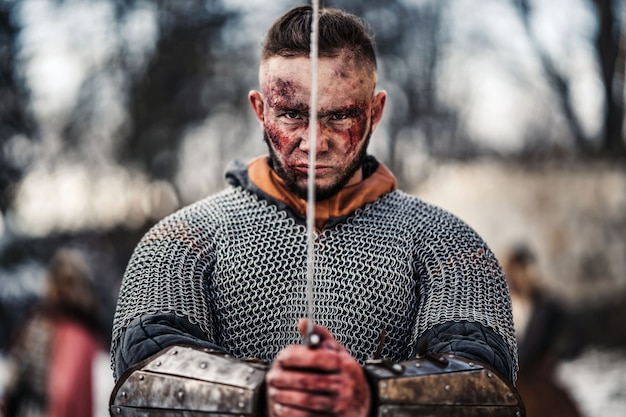 Il giovane uomo forte in un costume da guerriero medievale tiene una spada tra le mani proprio di fronte al suo viso. Concetto di guerra e storia