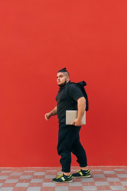 Il giovane uomo in sovrappeso in eleganti abiti neri cammina per strada con uno zaino sulla schiena