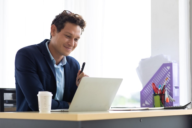 Il giovane uomo ispanico seduto beve caffè e lavora al computer portatile in ufficio creativo.