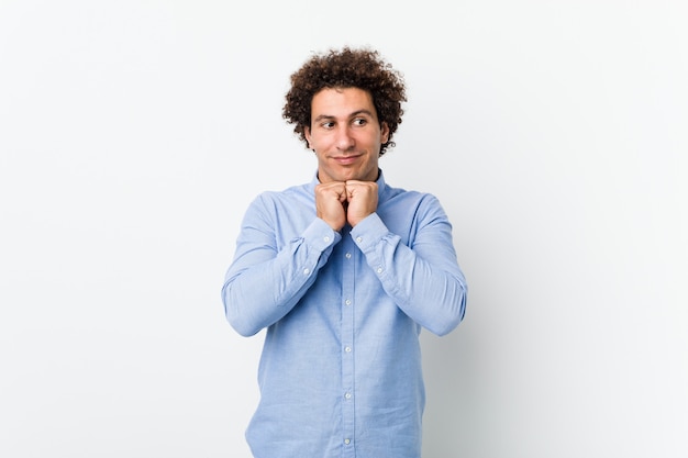 Il giovane uomo maturo riccio che indossa una camicia elegante tiene le mani sotto il mento, sta guardando felicemente da parte.