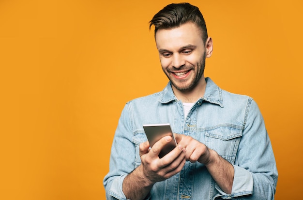 Il giovane uomo moderno ed elegante in denim sta usando lo smartphone nelle sue mani mentre è in piedi su sfondo giallo