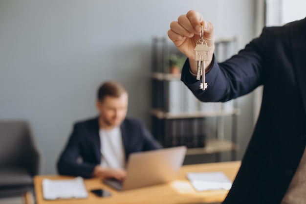 Il giovane uomo moderno sorridente in un vestito tiene le chiavi dell'appartamento acquistato sullo sfondo dell'agente immobiliare e dell'ufficio