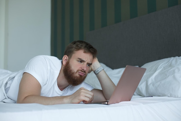 Il giovane uomo triste e infelice usa il tablet del gadget del computer portatile a casa a letto giaceva nella camera da letto guardando