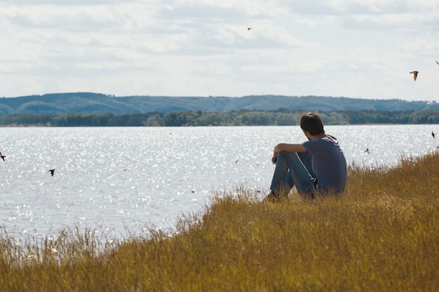 Il giovane uomo triste seduto sulla collina guarda in lontananza le rondini che volano sul fiume