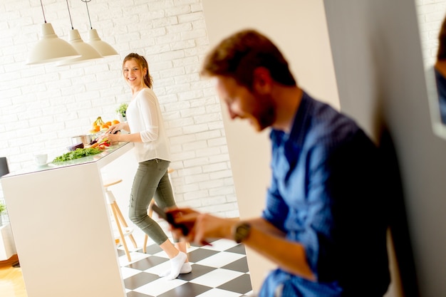 Il giovane usa un telefono cellulare nella cucina mentre la giovane donna cucina nei precedenti