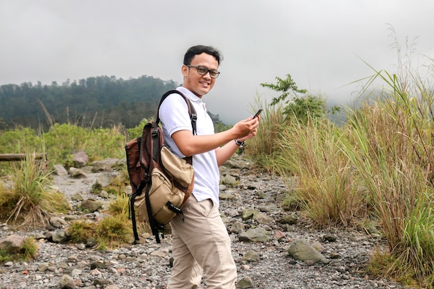 Il giovane viaggiatore con zaino e sacco a pelo asiatico è felice e sorride quando guarda lo smartphone quando viaggia in montagna