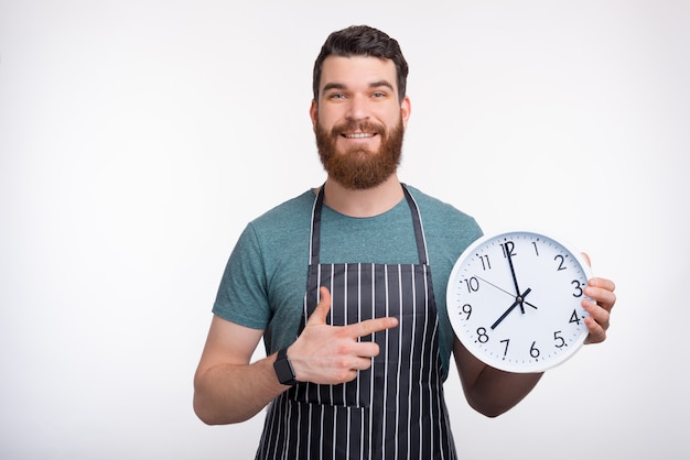 Il grembiule d'uso sorridente dell'uomo barbuto sta tenendo un orologio bianco mentre sorrideva sulla parete bianca