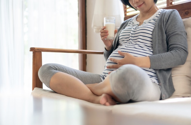 Il latte alimentare di seduta della giovane donna incinta allegra e tocca la sua pancia sul sofà a casa.