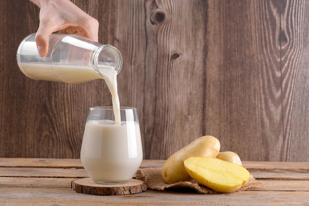 Il latte di patate vegano sta versando in un bicchiere e tuberi di patata su fondo di legno rustico a base di piante