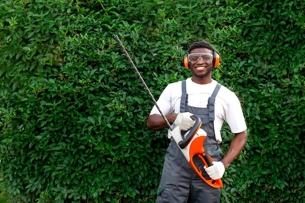 Il lavoratore del giardino in uniforme taglia i cespugli l'uomo afroamericano tiene il decespugliatore elettrico