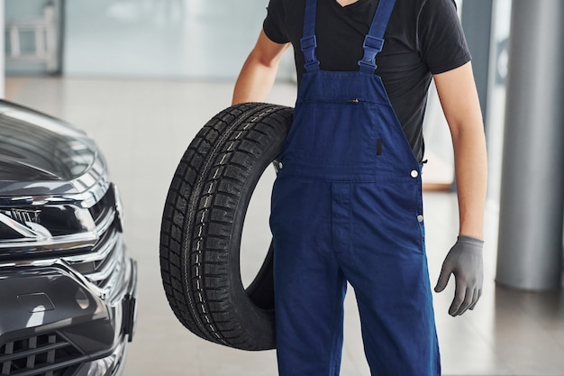 Il lavoratore in uniforme nera e blu che tiene la ruota di automobile e ha lavoro all'interno