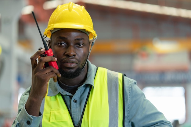 Il lavoratore nero maschio indossa il giubbotto di sicurezza con il casco giallo che tiene il walkietalkie mentre lavora in fabbrica