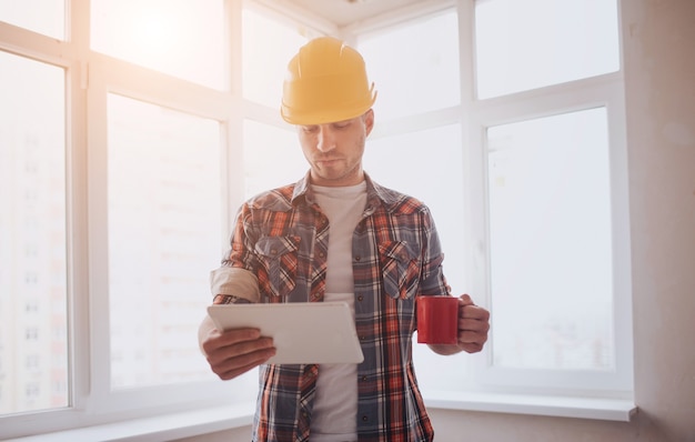 Il lavoratore o il costruttore tiene in mano una tazza di caffè e guarda il tablet. Sullo sfondo di costruzione e riparazione.