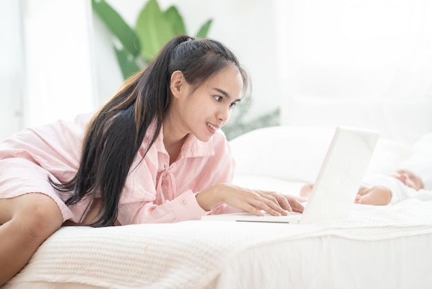 Il lavoro asiatico della giovane madre felice sul computer portatile sul letto ciao il concetto di cura neonata del sonno, del lavoro a casa e della scuola materna.