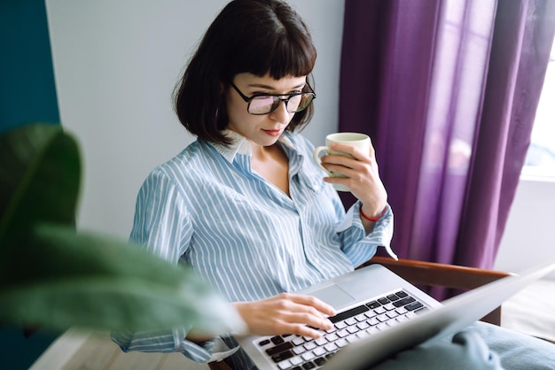 Il libero professionista della giovane donna si siede sul sofà moderno e lavora sul computer portatile. Istruzione in linea. Lavorare da casa.