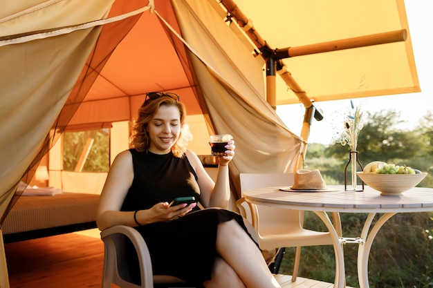 Il libero professionista Happy Woman usa il telefono e beve cocktail mentre si rilassa su un'accogliente tenda glamping in una giornata di sole Tenda da campeggio di lusso per vacanze estive all'aperto e vacanze Concetto di stile di vita