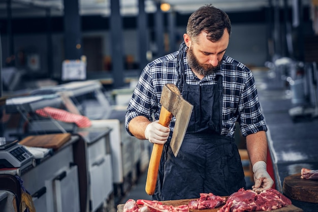 Il macellaio barbuto vestito con una camicia di pile taglia la carne con un'ascia insanguinata.