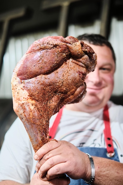 Il macellaio tiene un cosciotto di agnello condito con la marinata davanti a un grande fumatore