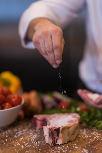 Il maestro chef mette sale su una succosa fetta di bistecca cruda con verdure su un tavolo di legno