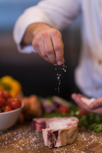 Il maestro chef mette sale su una succosa fetta di bistecca cruda con verdure su un tavolo di legno