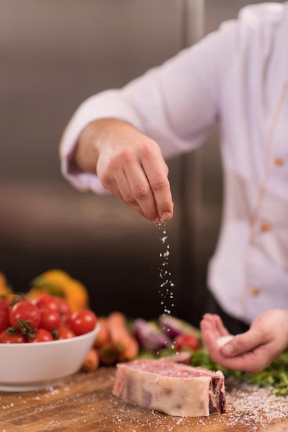 Il maestro chef mette sale su una succosa fetta di bistecca cruda con verdure su un tavolo di legno