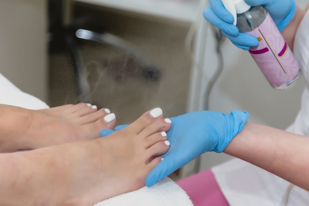 Il maestro di pedicure in guanti di gomma blu spruzza i piedi del cliente prima della procedura di cura delle unghie dei piedi. Avvicinamento. Pedicure nel salone di bellezza. Igiene e cura dei piedi. Concetto di industria della bellezza