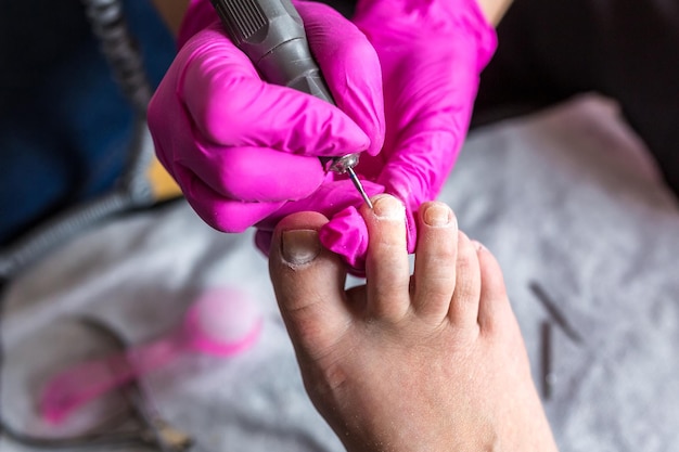 Il maestro pedicure in guanti rosa taglia le unghie dei piedi della cuticola e della gommalacca nel salone di pedicure usando il trapano Pedicure professionale nella clinica di cosmetologia Igiene dei piedi nel salone di bellezza