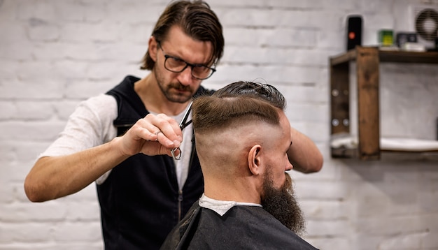 Il maestro taglia i capelli e la barba degli uomini nel barbiere, il parrucchiere fa acconciatura per un giovane