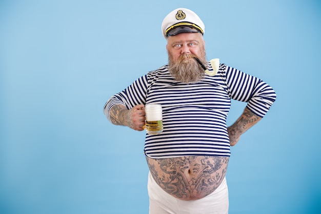 Il marinaio grasso e focoso con la barba e la pipa fumante tiene la birra schiumosa su sfondo azzurro