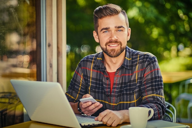 Il maschio barbuto bello beve il caffè mentre usa un computer portatile in un caffè.