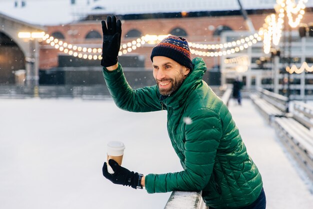 Il maschio barbuto sorridente beve il caffè asportabile, ondeggia con la mano il suo amico, ha un'espressione allegra